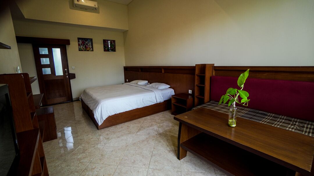 A bedroom with a bed, a wooden nightstand, a wooden table with a plant, a red sofa, and a door with a mirror.
