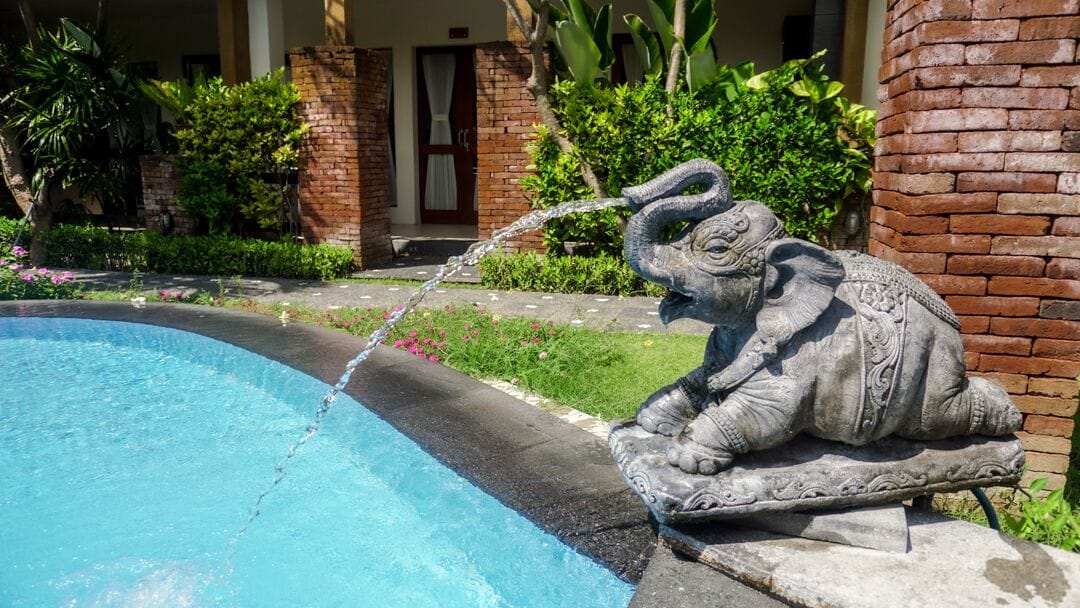 Uma estátua de uma figura sentada perto de uma piscina, com um muro de tijolos e vegetação ao fundo.