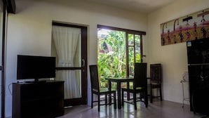 Quarto com televisão em suporte, mesa com cadeiras, porta para varanda com plantas e frigorífico.
