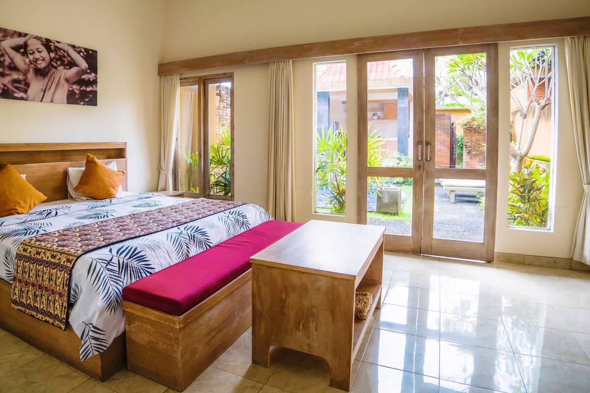 A hotel room with a large bed, a wooden bench, and a view of the garden through the glass doors.