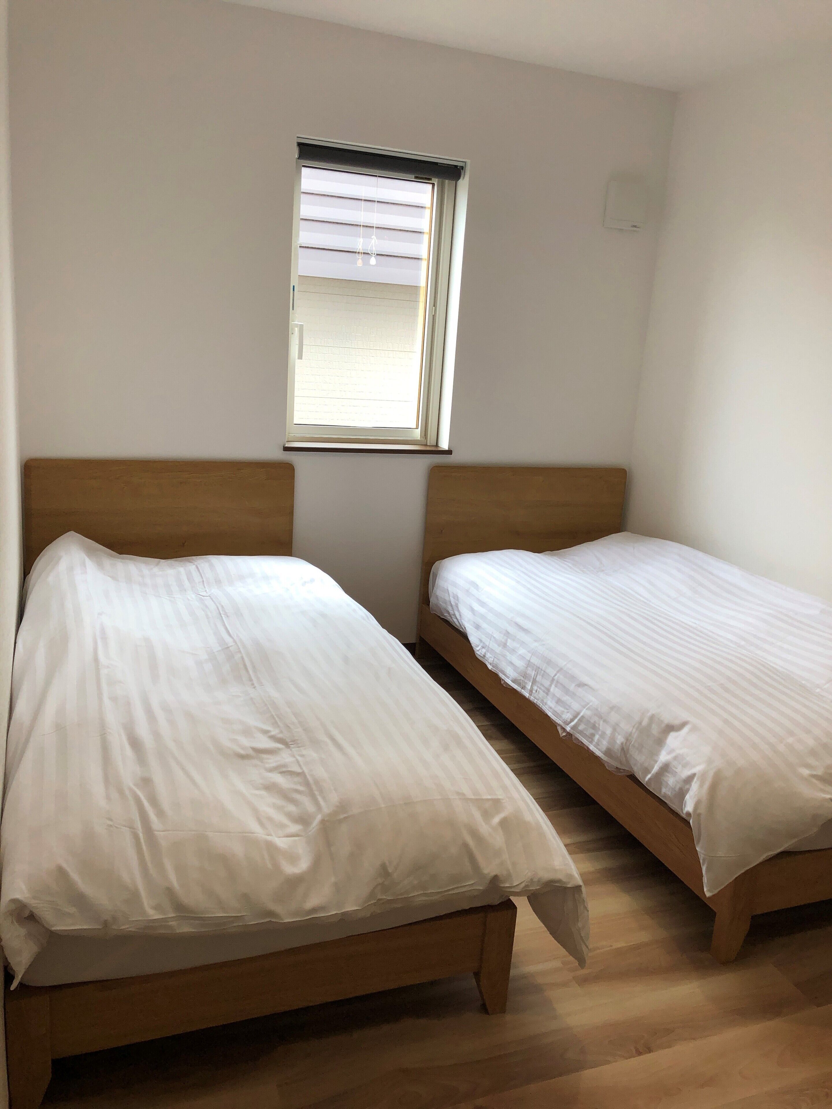 Een kamer met twee houten bedden, een raam met zonwering en een lichtschakelaar aan de muur.