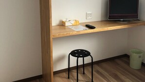 Een kleine kamer met een houten bureau, een televisie, een klein krukje en een afvalbak.
