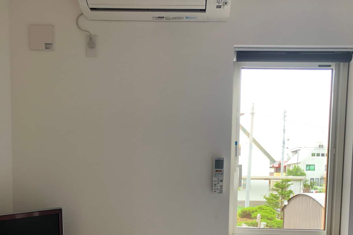 A hotel room with a bed, a TV on a wooden shelf, a window with a view of buildings, and an air conditioning unit.