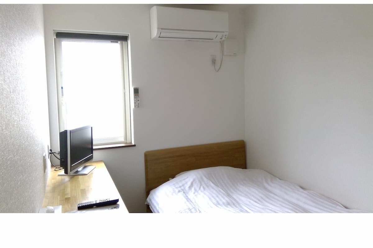 A small, sparsely furnished room with a bed, a desk, a television, and a window.