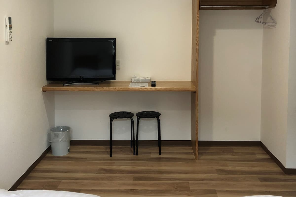 A hotel room with a bed, a TV on a wooden shelf, two black stools, a trash bin, and a wooden wardrobe.