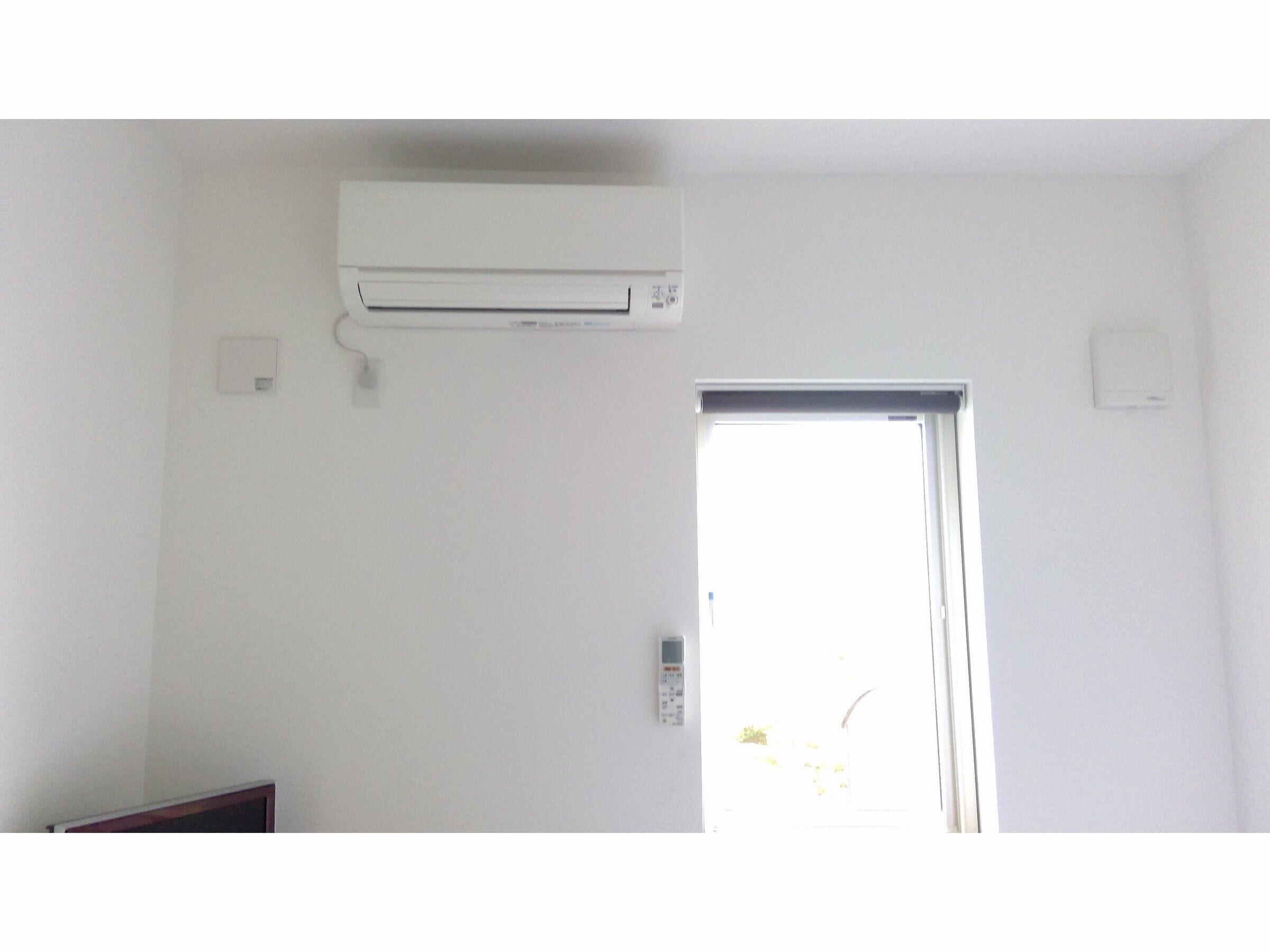 Una habitación con pared blanca, aire acondicionado instalado en el techo, una puerta con ventanal y un pequeño enchufe eléctrico.