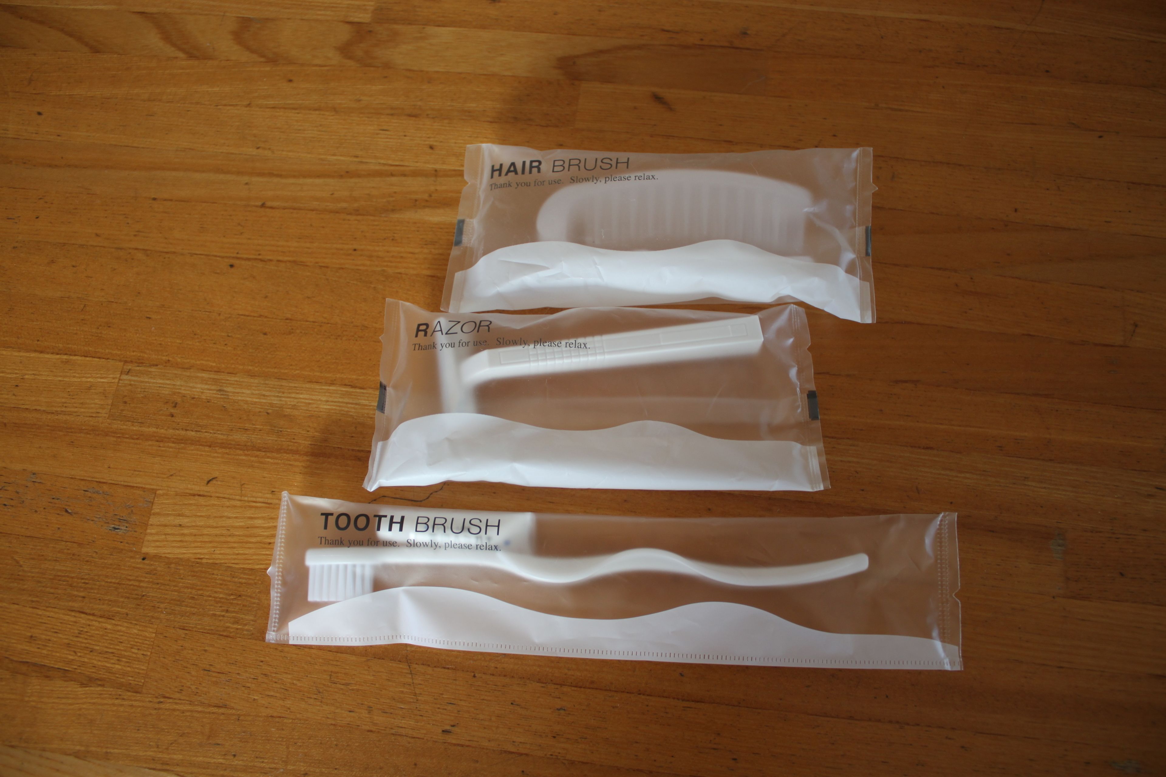 Three white plastic packages labeled "HAIR BRUSH," "RAZOR," and "TOOTH BRUSH" on a wooden floor.