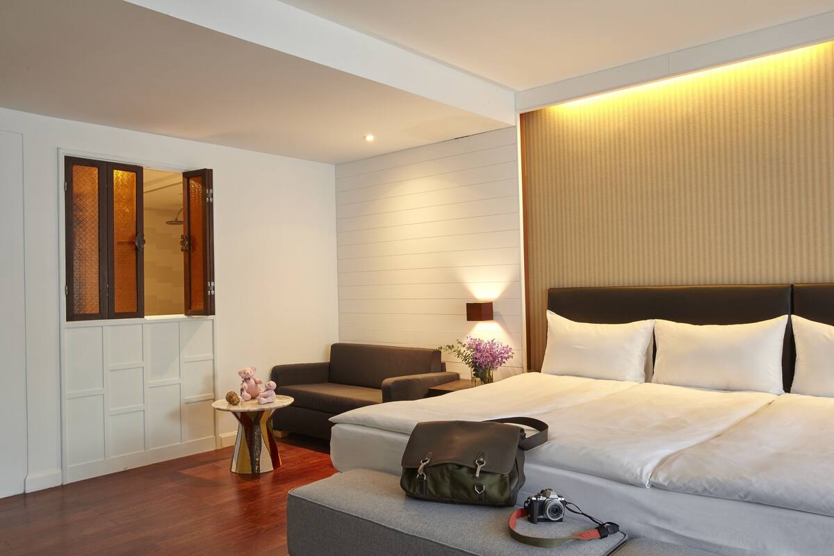 A hotel room with a bed, a sofa, a small table with a vase of flowers, and a camera on the ottoman.