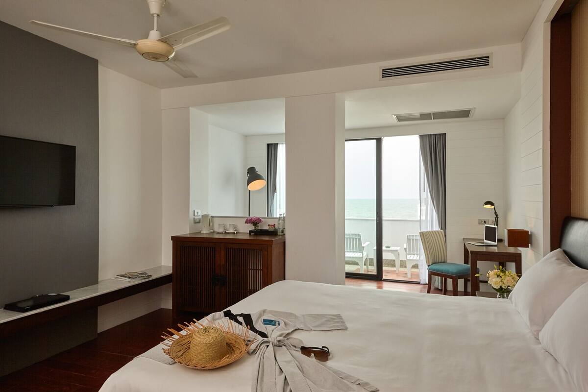 A hotel room with a bed, a desk, a chair, a TV, and a balcony with a view of the sea.