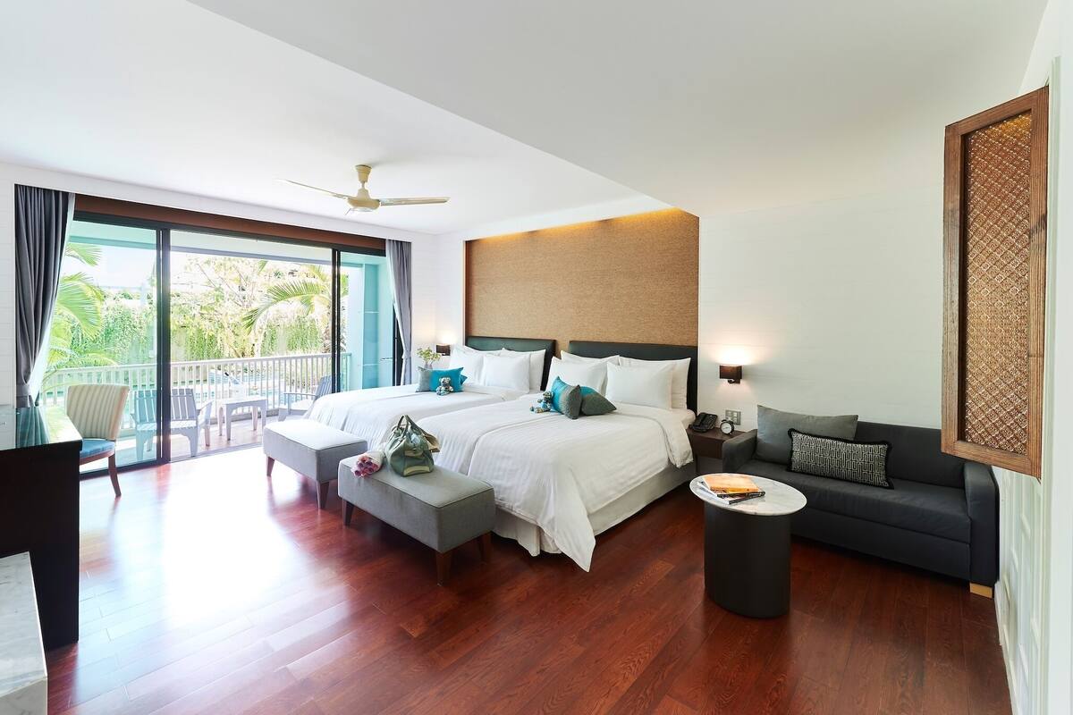 A hotel room with a large bed, a sofa, a small table, a desk, and a balcony with chairs.