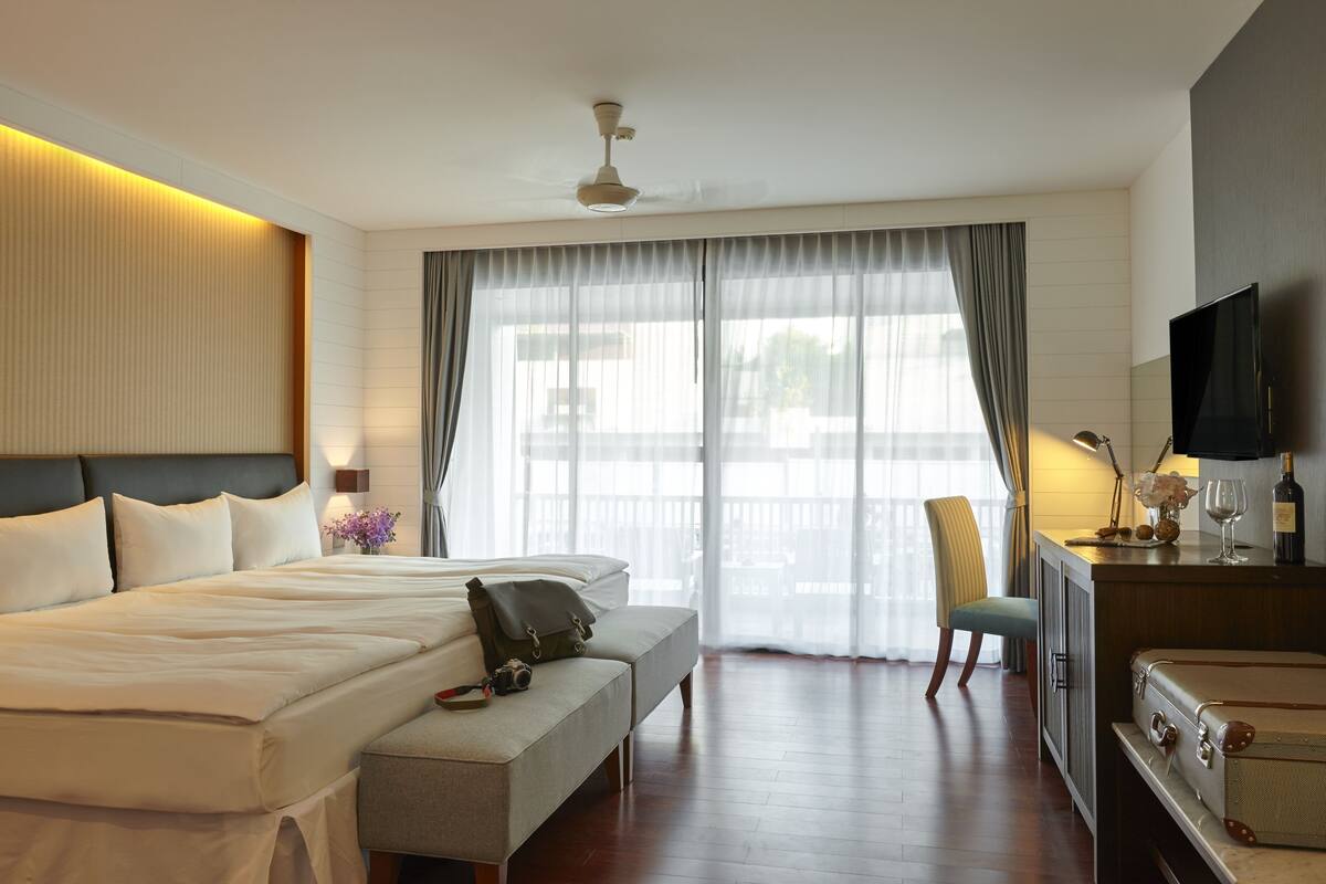 A hotel room with a large bed, a desk with a flat-screen TV, a chair, and a bench.