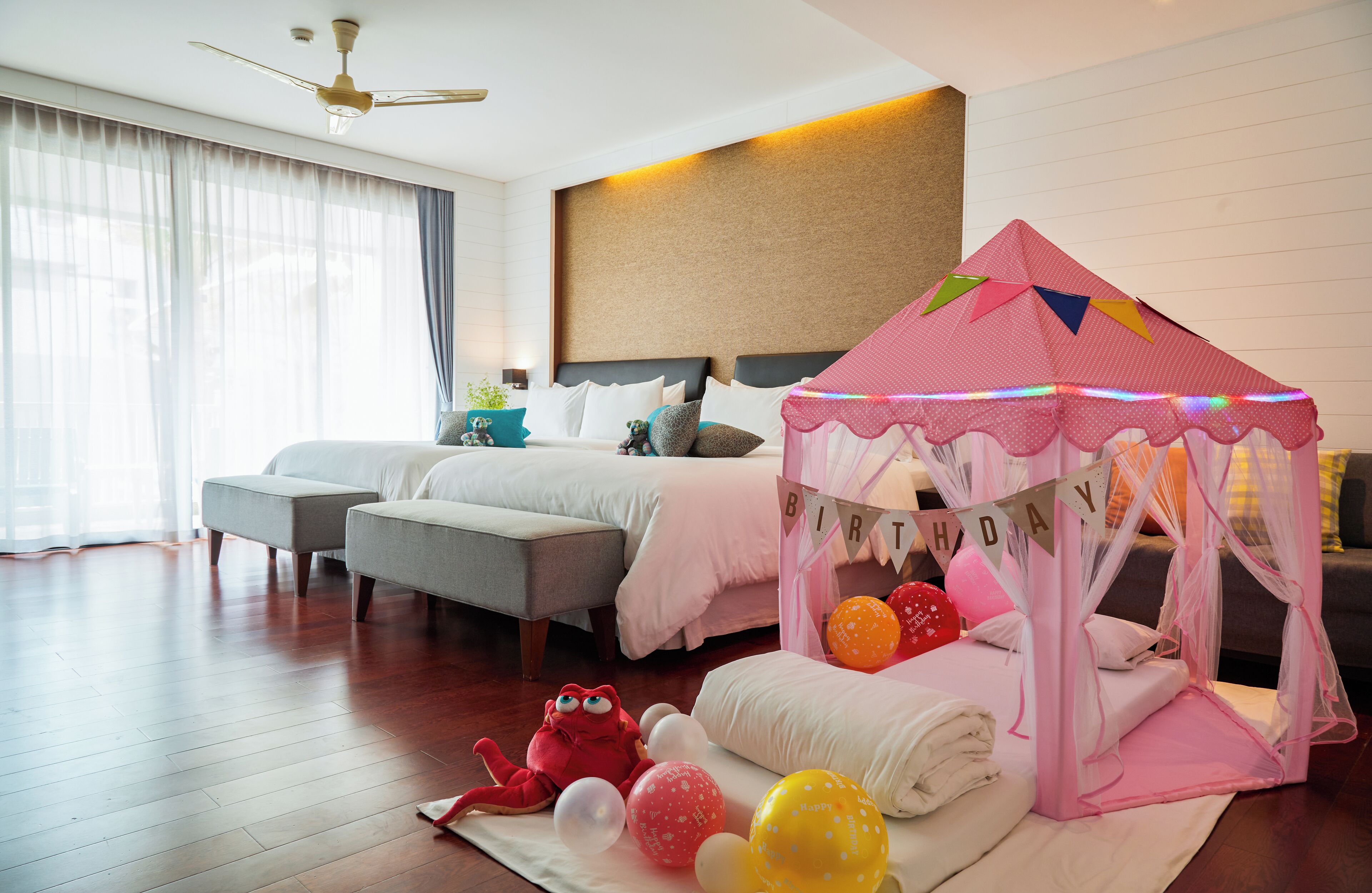 A bedroom with two beds, a ceiling fan, a tent with 'BIRTHDAY' banner, and balloons.