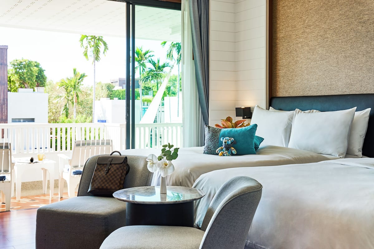 A hotel room with a balcony, a bed with white linens, a small round table with a vase of flowers, and a grey armchair.