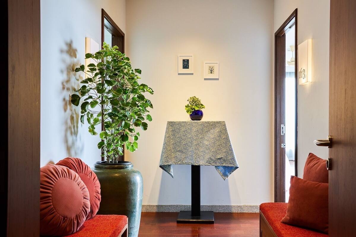 A hallway with a large potted plant, a small table with a patterned tablecloth, and two red cushioned chairs.