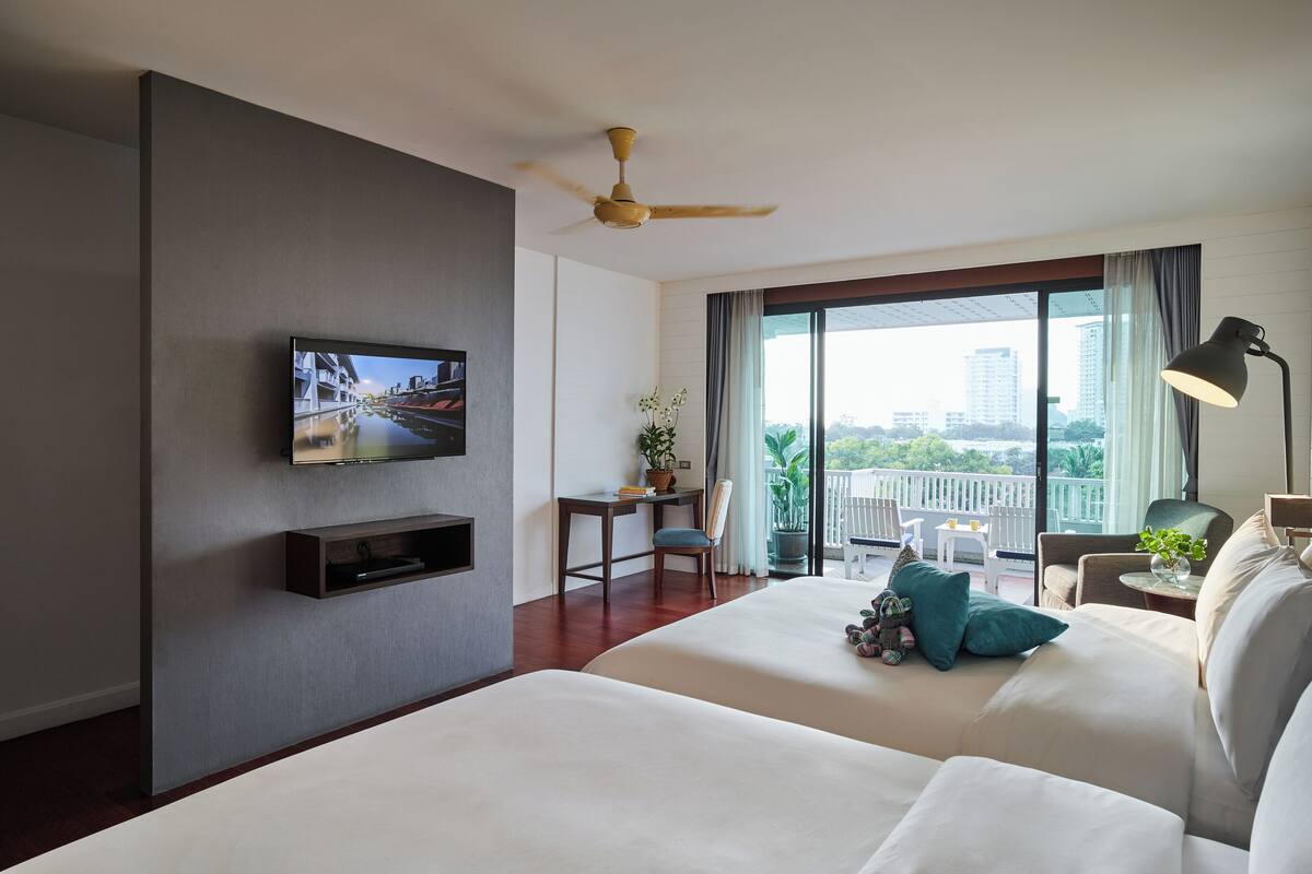 A hotel room with two beds, a desk, a TV, a balcony with a view of the city, and a ceiling fan.