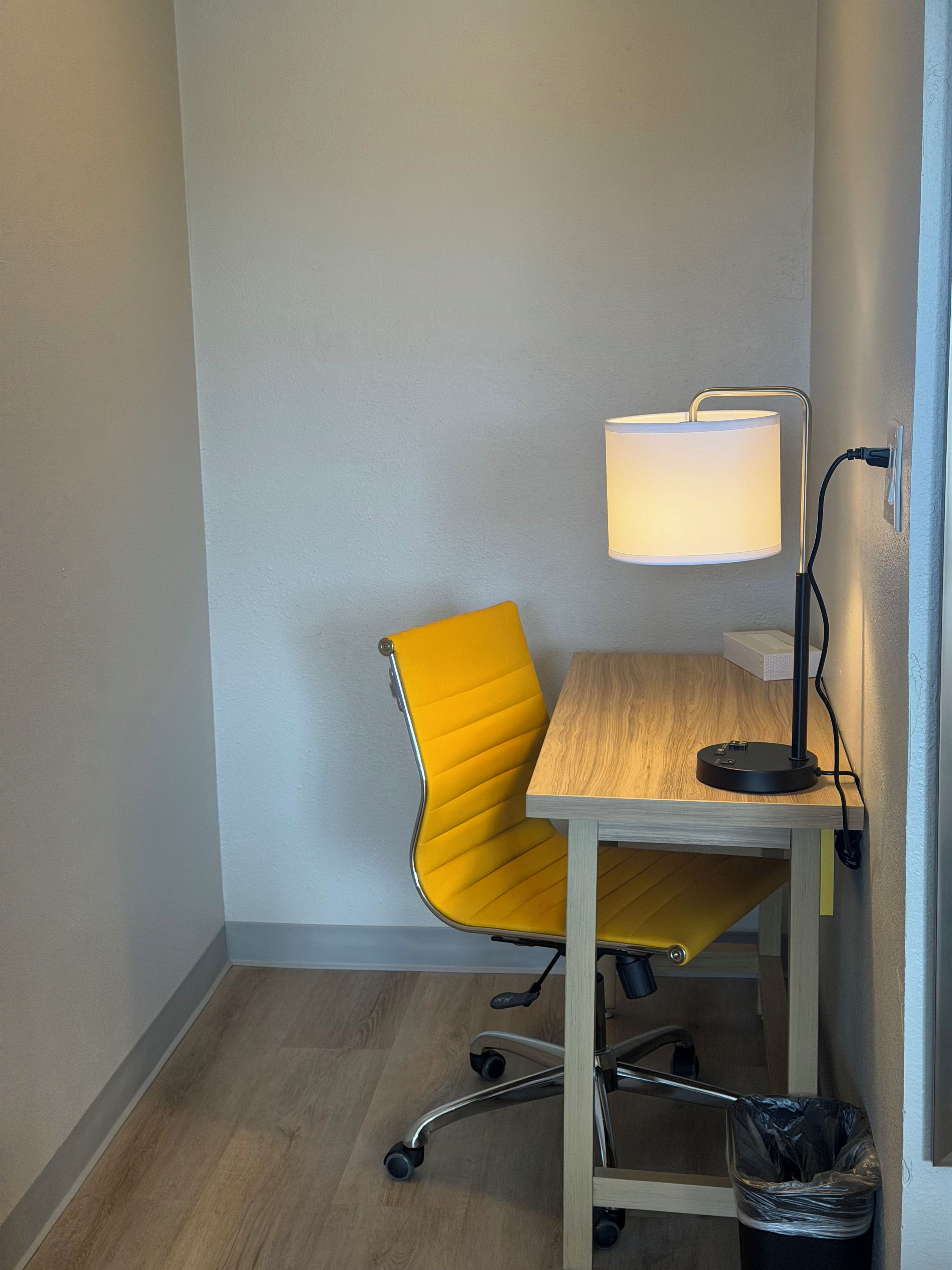 Une pièce d’angle avec une chaise jaune, une table en bois, une lampe de bureau et une poubelle.