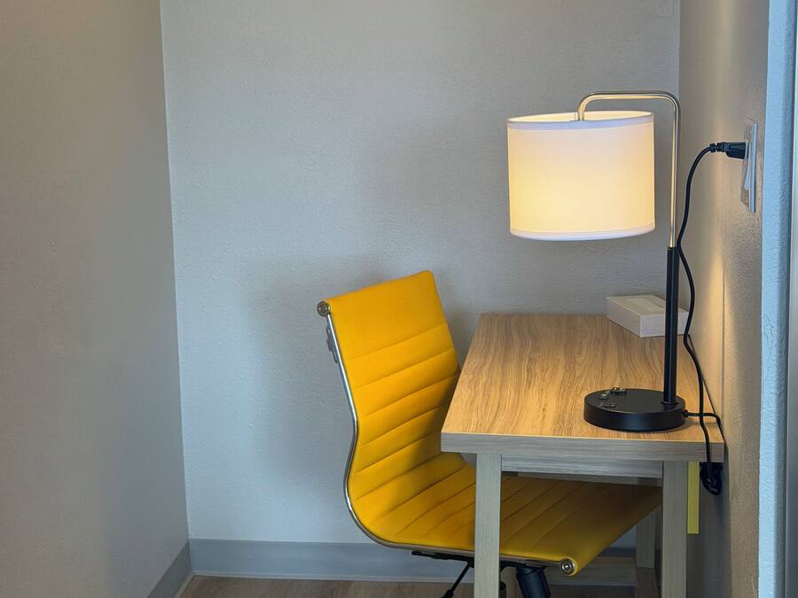 Una habitación de esquina con una silla amarilla, una mesa de madera, una lámpara de escritorio y un cesto de basura.