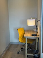 Una habitación de esquina con una silla amarilla, una mesa de madera, una lámpara de escritorio y un cesto de basura.