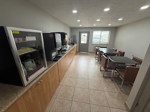 A clean, modern kitchen area with a refrigerator, microwave, and coffee machine. There is a dining table with chairs and a window providing natural light.