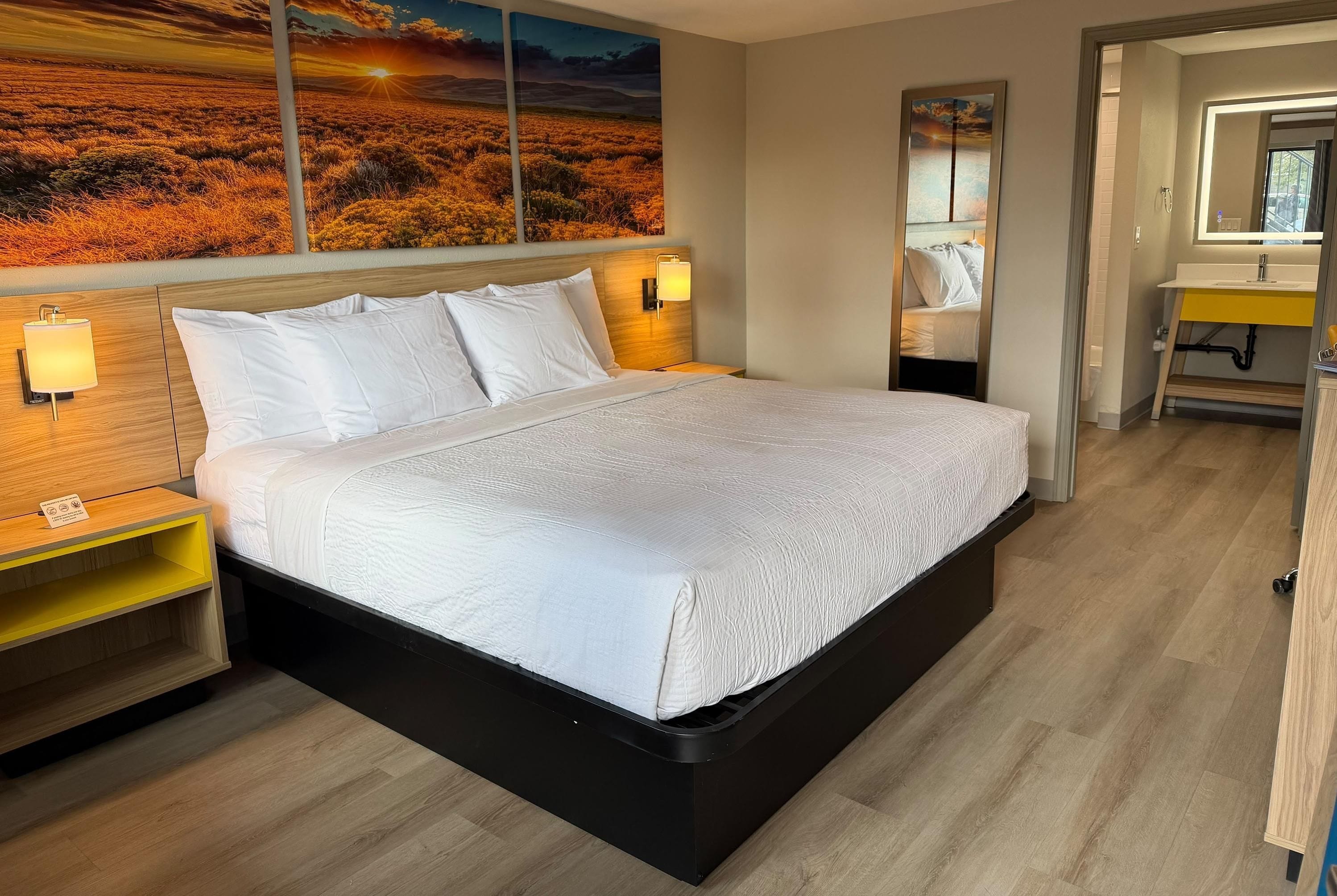 Moderni hotellihuone, jossa on suuri sänky, yöpöytävalaisimet, puinen pääty ja maalaus, jossa on kuvattu auringonlaskun maisema.