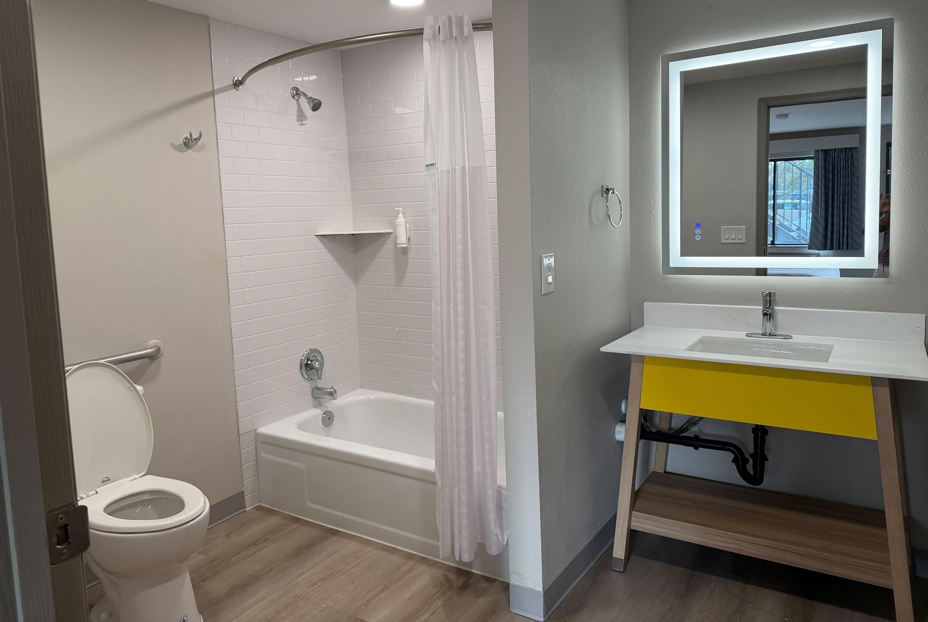A bathroom with a white toilet, a bathtub, a shower with a curtain, a sink with a mirror, and a window.