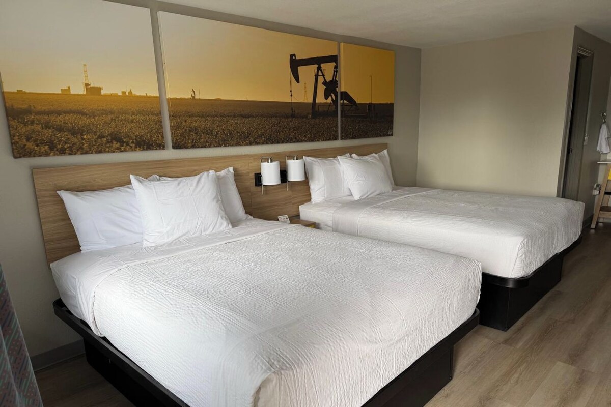 Une chambre d’hôtel avec deux lits, une grande peinture représentant un champ pétrolier et une tête de lit en bois.