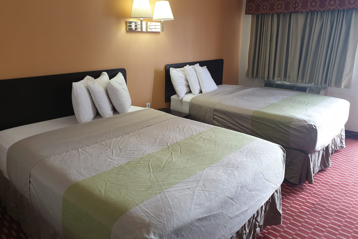 A hotel room with two beds, each with white bedding and a green accent. There are wall-mounted lamps above the beds and a window with curtains.