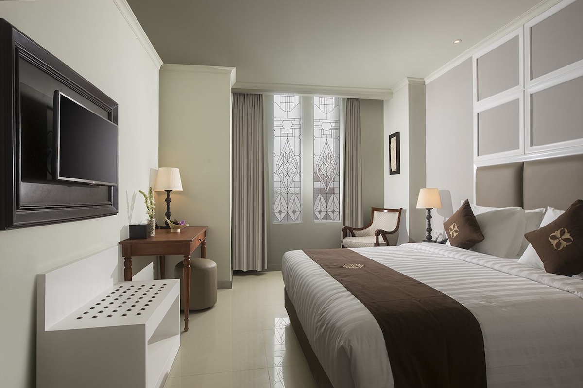 A hotel room with a bed, bedside tables, a TV, a window with curtains, and a wall-mounted TV.