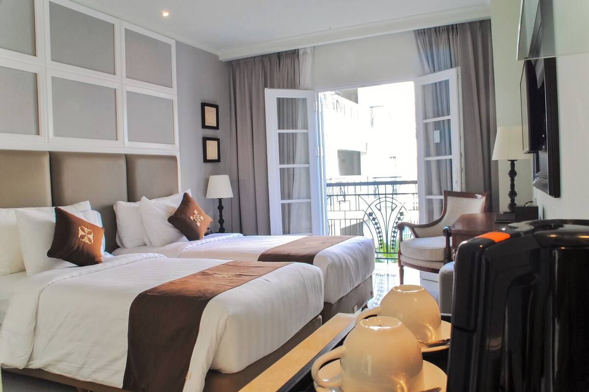 A hotel room with two beds, a balcony, a chair, a table with a lamp, and a TV.