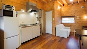 Cocina compacta con microondas, estufa y lavamanos. Hay una silla blanca y una pequeña mesa. La habitación tiene paredes y techo de madera.