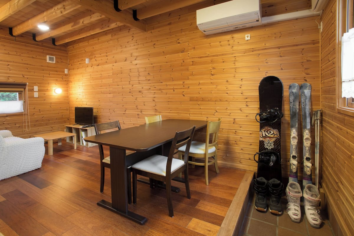 A wooden cabin interior with a dining table, chairs, a sofa, and skis and ski boots against the wall.