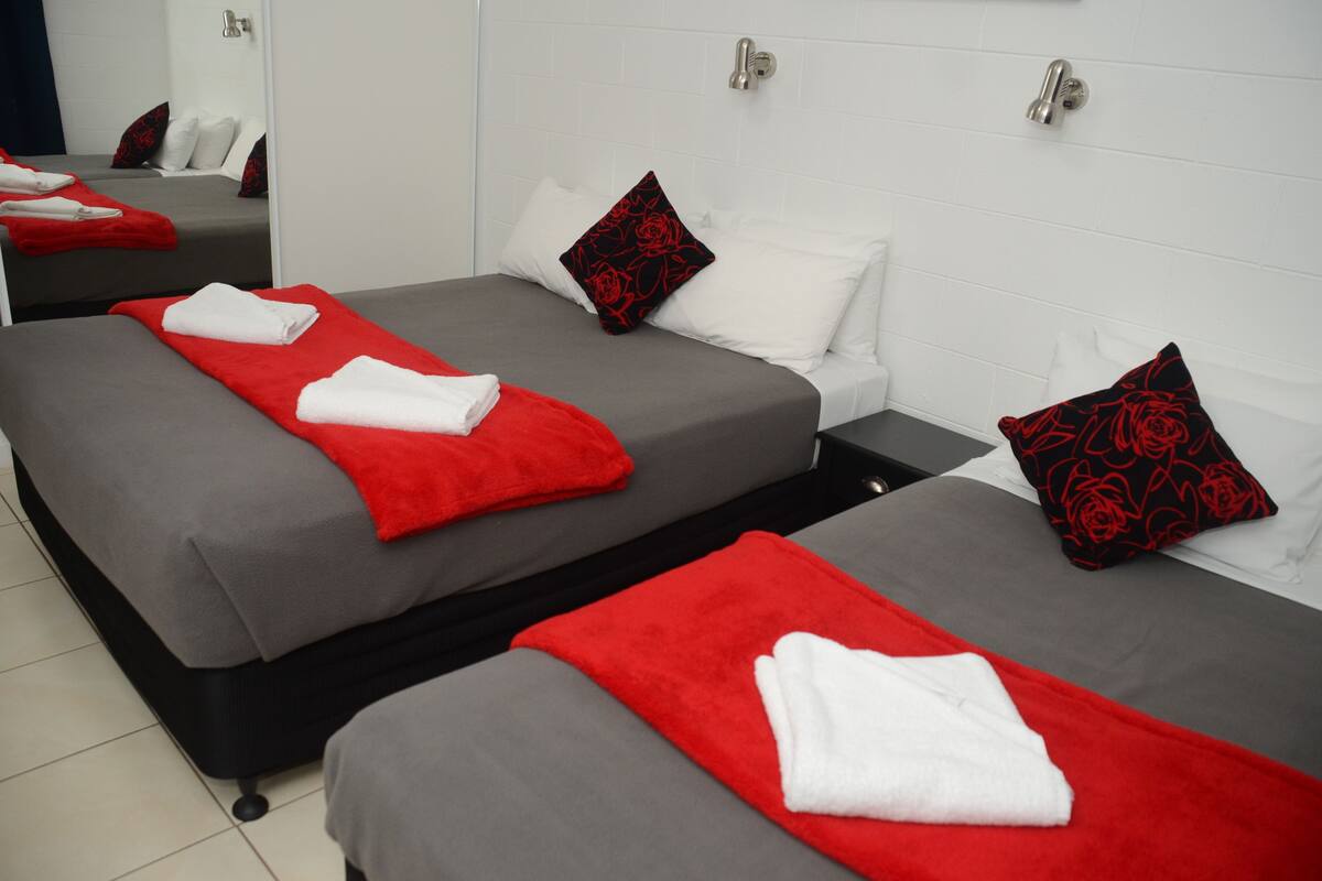 A hotel room with three beds, each with a red and white blanket, and red rose-patterned pillows.