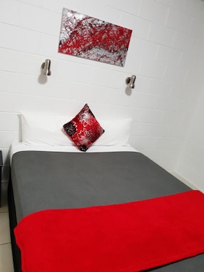 Ein Hotelzimmer mit Bett, weißen Wänden, einem rot-schwarzen Kopfkissen und einer roten und grauen Decke.