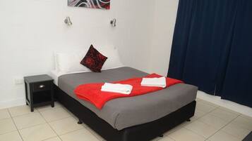 Una cama bien hecha con una manta gris y roja, una mesita de noche negra, un almohadón blanco y un almohadón rojo con diseño floral.