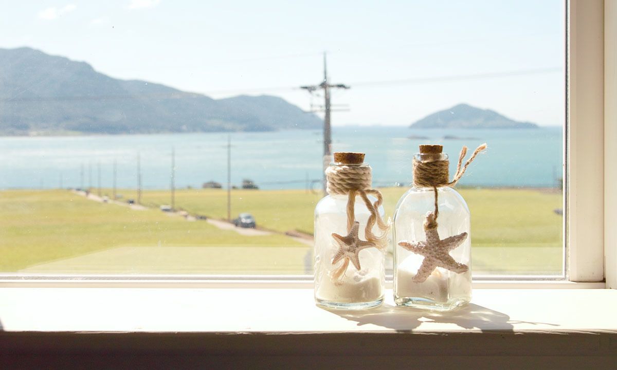 Dos botellas de vidrio con estrellas de mar y cuerda, sobre un alféizar con vistas a una masa de agua y montañas lejanas.