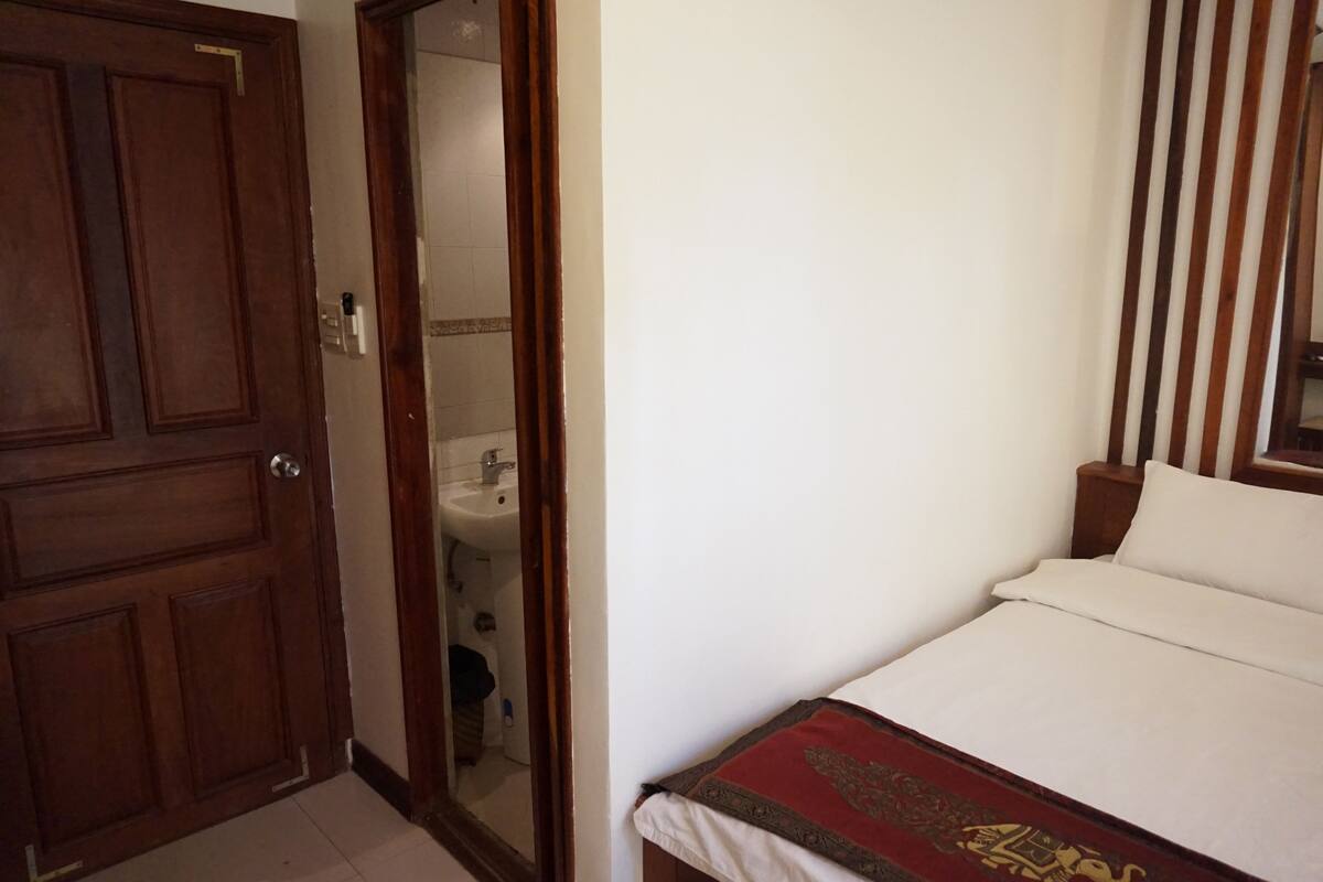 A bedroom with a wooden door, a white bed with a red and gold bedspread, and a wooden headboard.