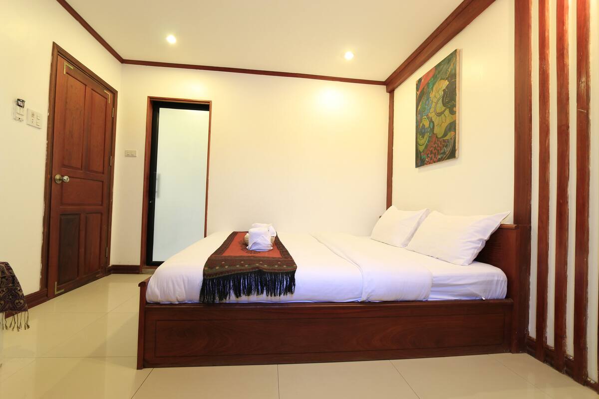 A bedroom with a wooden bed, white bedding, a red and black throw blanket, and a painting on the wall.