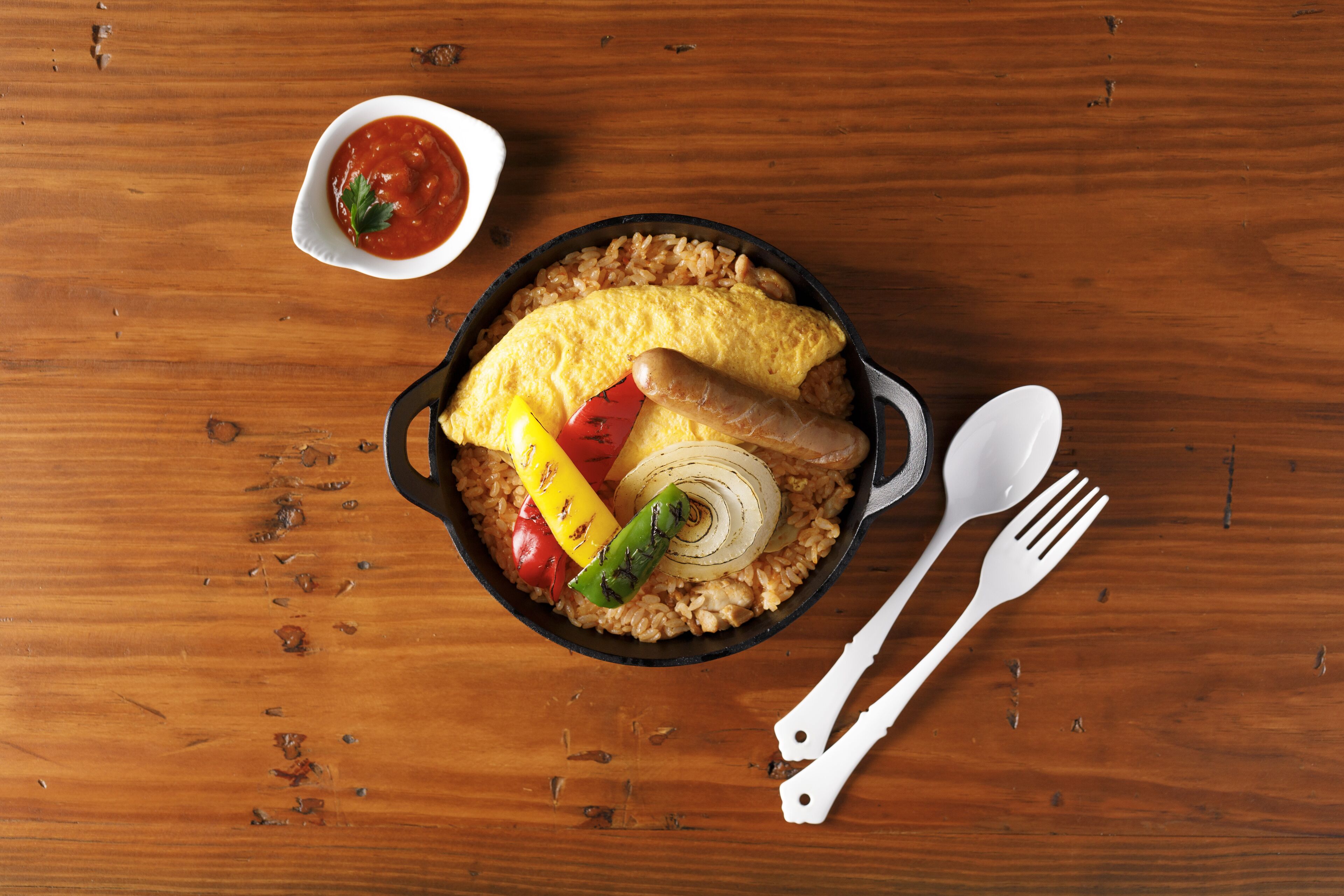 A meal with rice, vegetables, and an omelette in a skillet, accompanied by a small bowl of sauce and white utensils on a wooden table.