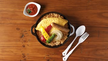 A meal with rice, vegetables, and an omelette in a skillet, accompanied by a small bowl of sauce and white utensils on a wooden table.