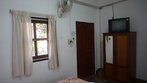 Una habitación pequeña con cama, un armario de madera, un televisor encima del armario, una ventana con cortinas y un ventilador fijado a la pared.