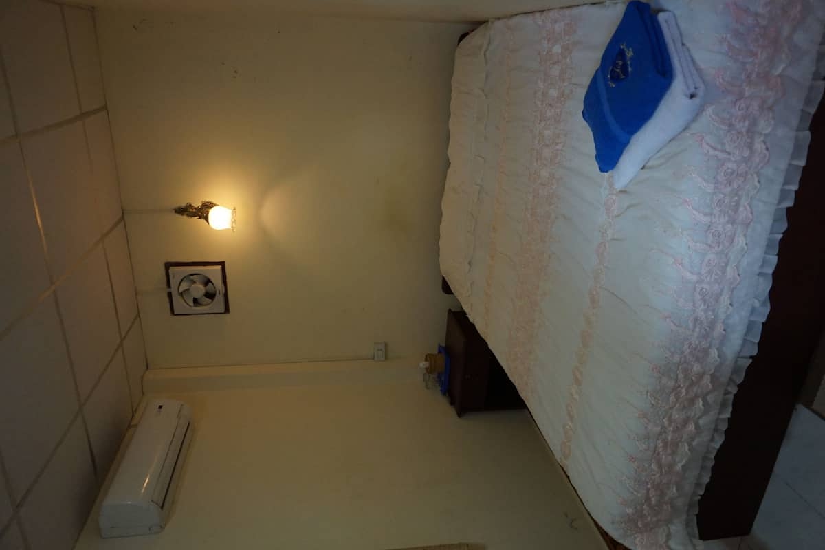 A small, dimly lit room with a single bed, a wall-mounted air conditioner, and a small shelf with a blue item.