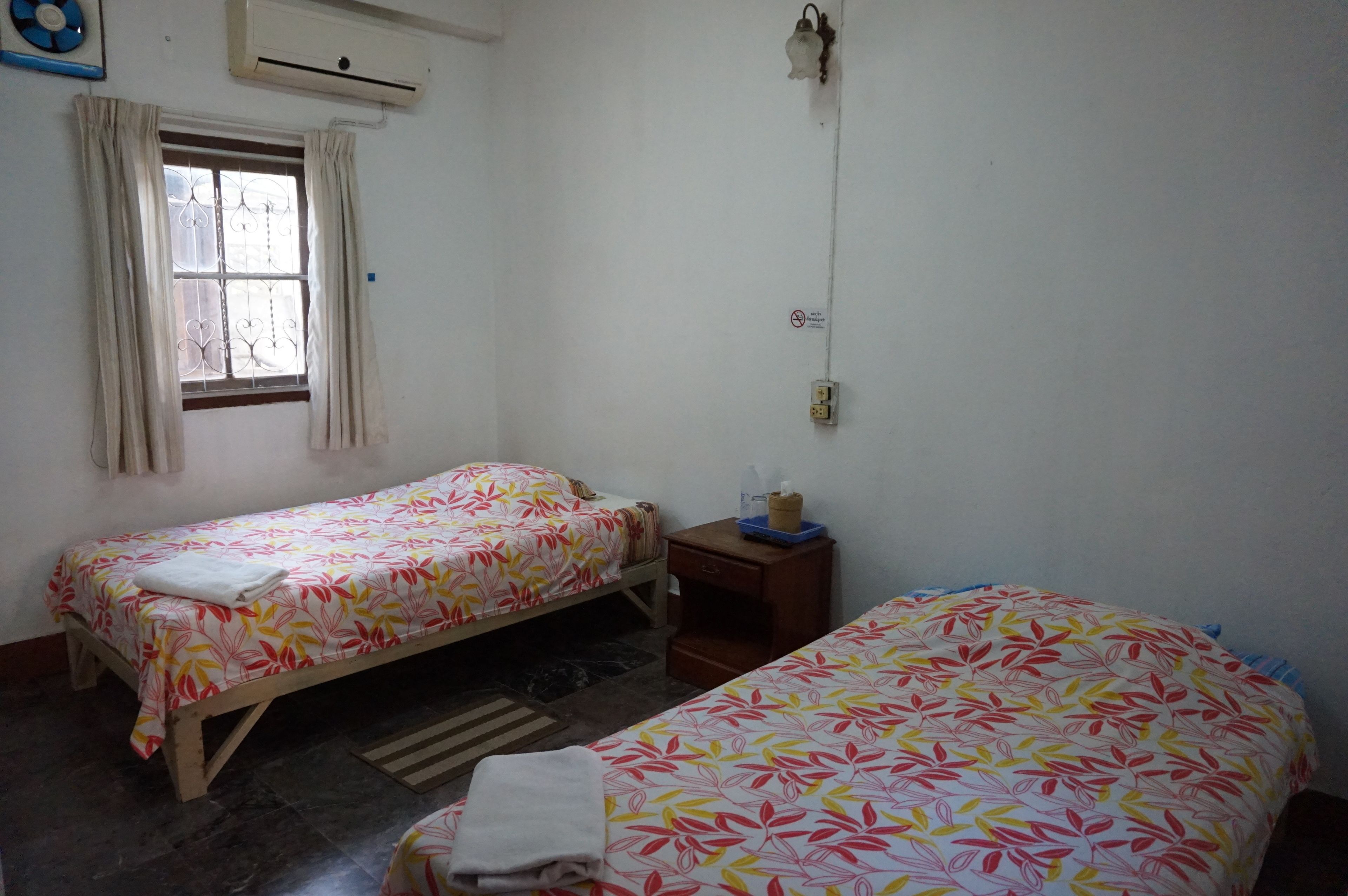 Una habitación pequeña y limpia con dos camas individuales, una ventana con cortinas, una mesita de noche con lámpara y un equipo de aire acondicionado.