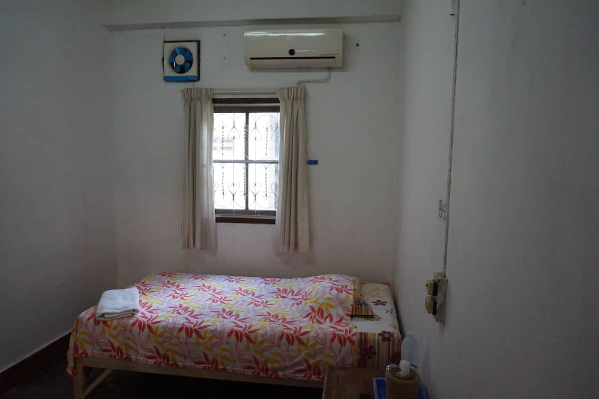 A small, simple room with a bed, a window with curtains, an air conditioner, and a wall clock.