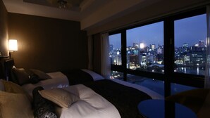 Een hotelkamer met een groot bed, een klein rond tafeltje en uitzicht op een stadsbeeld in de schemering.