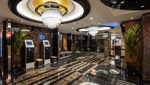 Een moderne hotel lobby met een marmeren vloer, een grote kroonluchter en een receptiebalie met zelfbedieningskiosken.