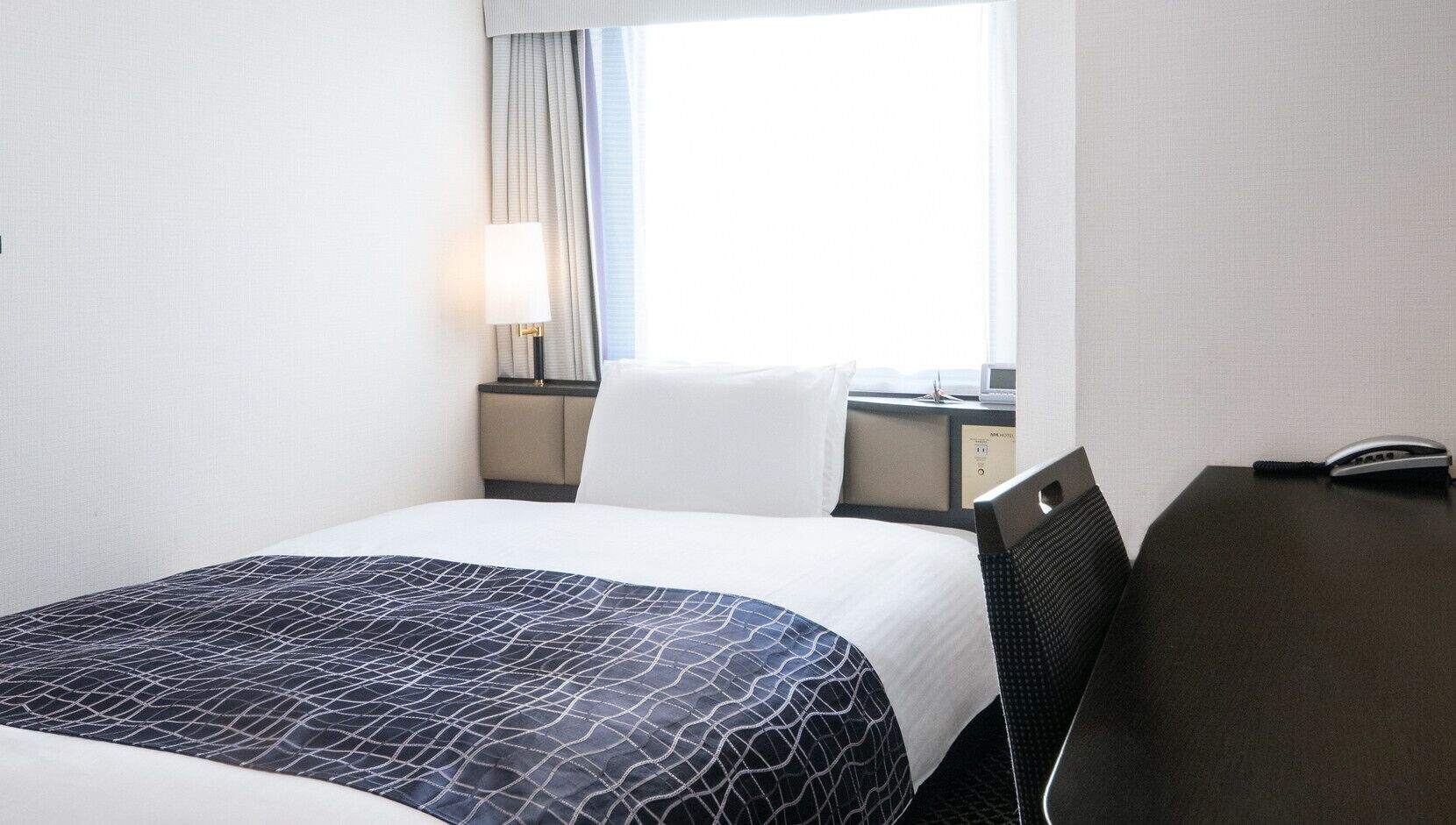 Une chambre d’hôtel avec un lit, un bureau, une lampe, une chaise et une fenêtre avec des rideaux.