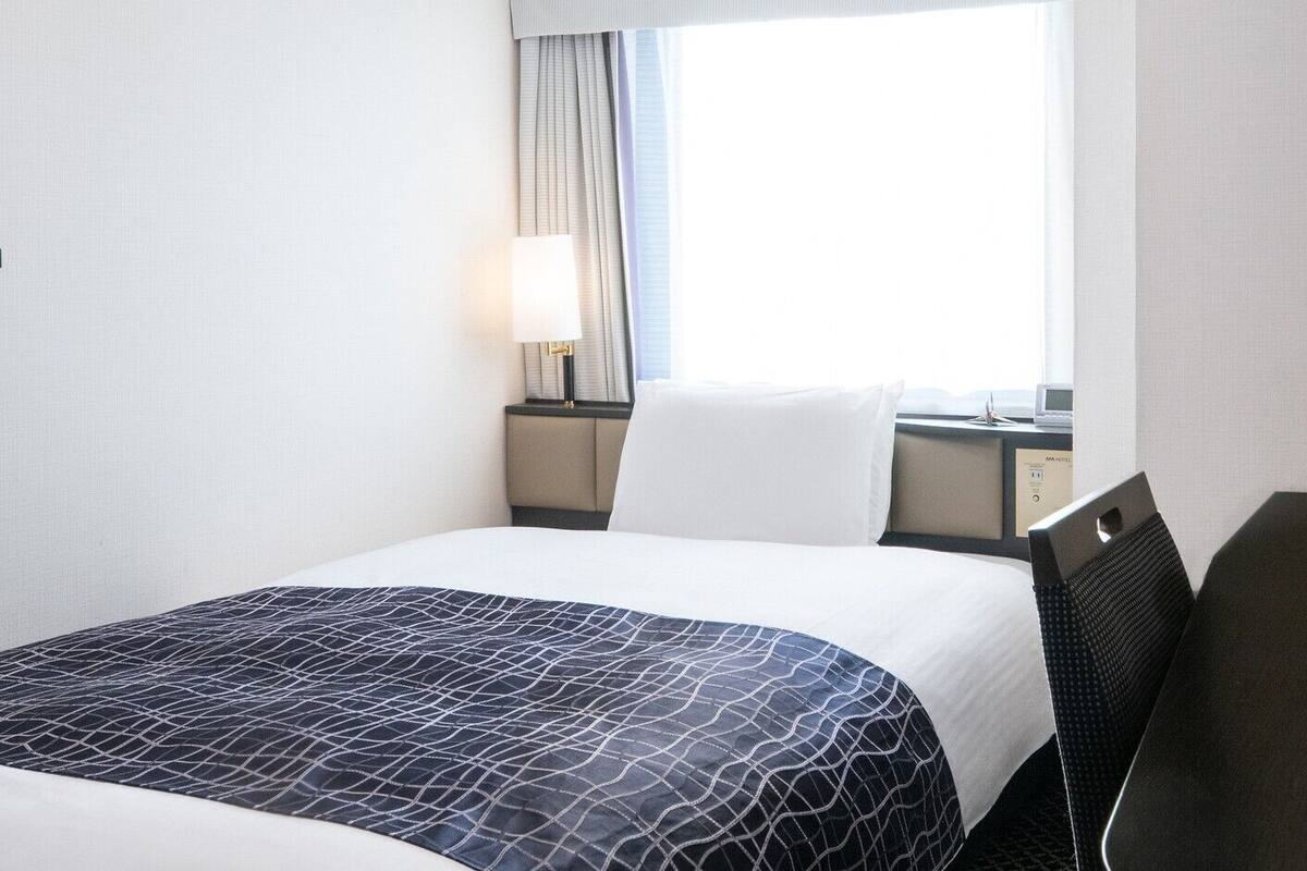 Une chambre d’hôtel avec un lit, un bureau, une lampe, une chaise et une fenêtre avec des rideaux.