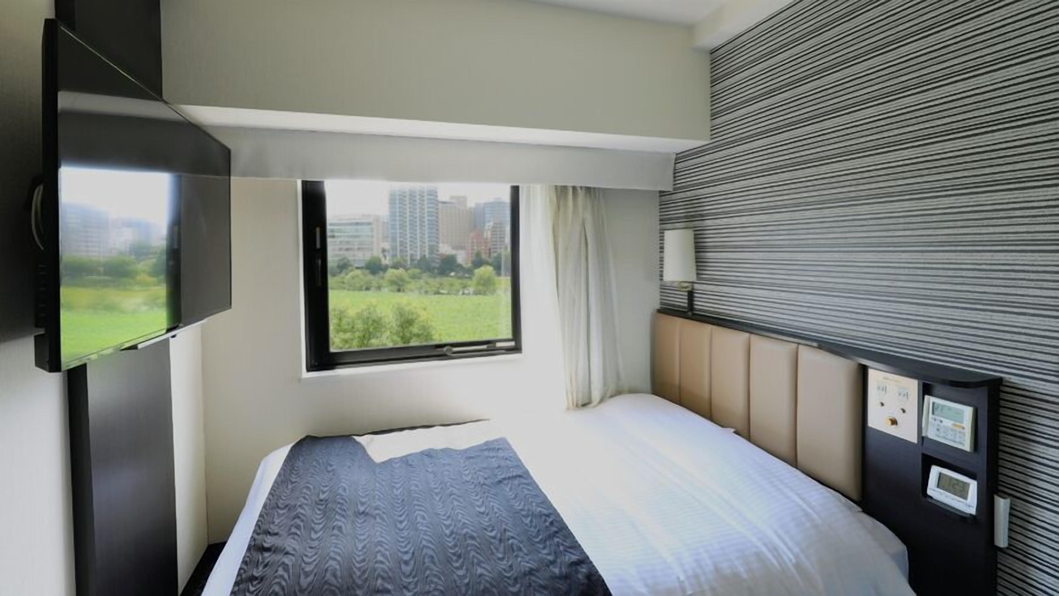 Une chambre d’hôtel moderne équipée d’un lit, d’un téléviseur fixé au mur, d’une fenêtre donnant sur un paysage verdoyant et des bâtiments, ainsi que d’un panneau de commande mural.