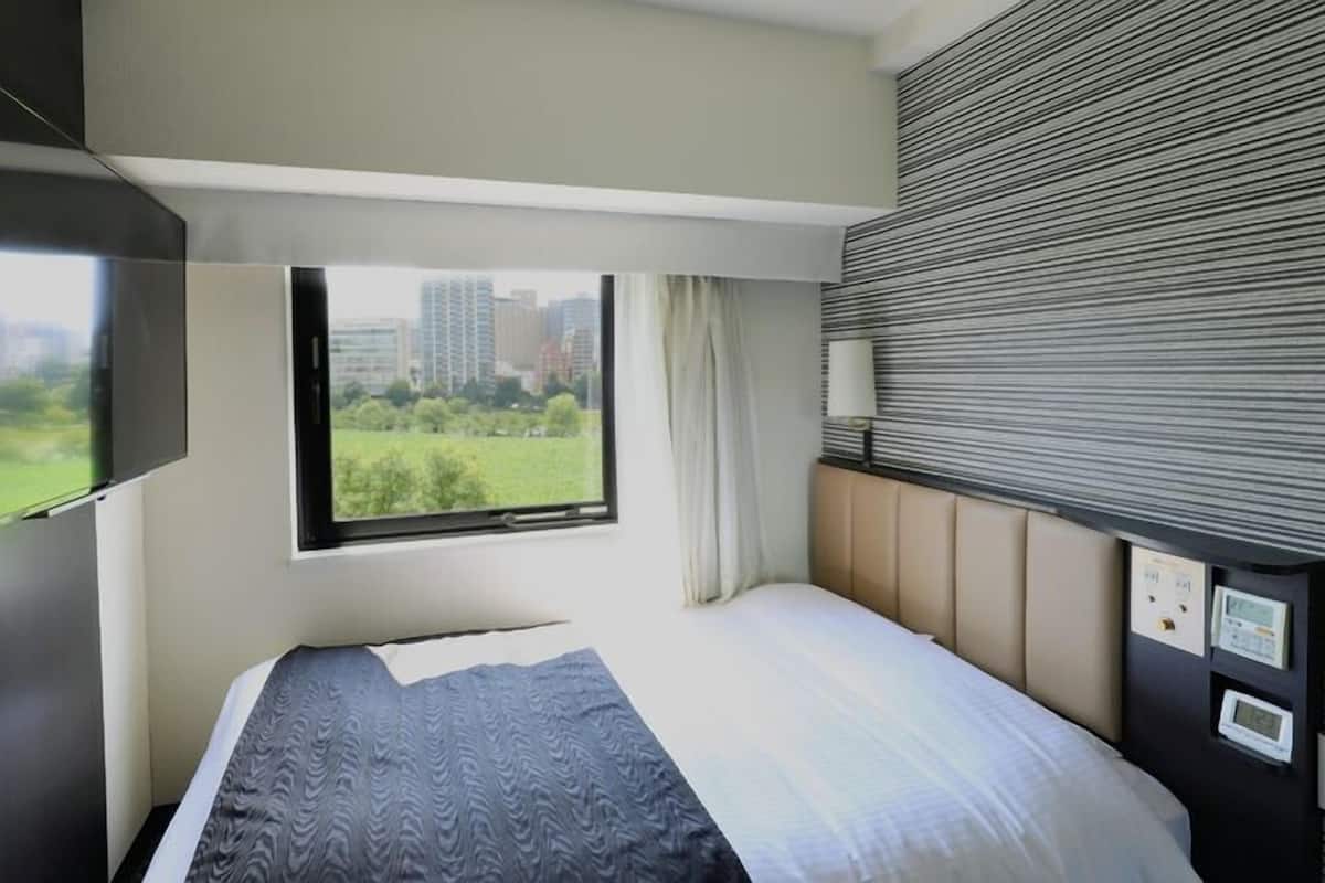 Een moderne hotelkamer met een bed, een aan de muur bevestigde televisie, een raam met uitzicht op groen en gebouwen, en een bedieningspaneel aan de muur.