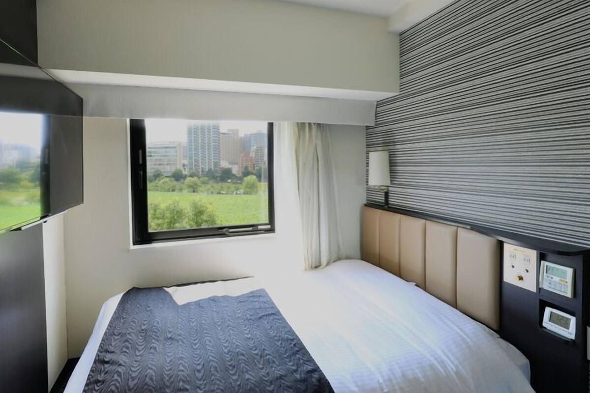 Une chambre d’hôtel moderne équipée d’un lit, d’un téléviseur fixé au mur, d’une fenêtre donnant sur un paysage verdoyant et des bâtiments, ainsi que d’un panneau de commande mural.